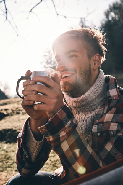 Man sitting outdoors holding a mug, wearing a plaid shirt and sweater.