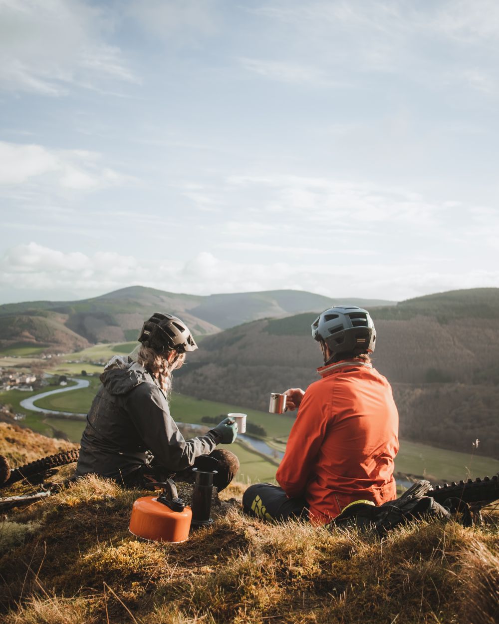 Two people sitting on a hilltop with scenic views and mountains in the background.