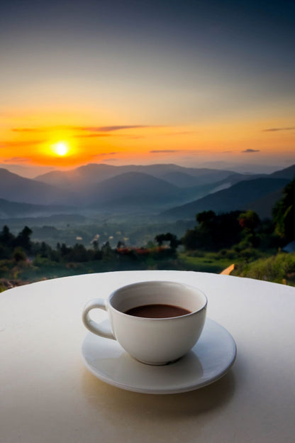 White cup of coffee on a saucer with a scenic sunset over mountains in the background